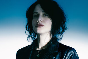 Portraitfoto von Hüma Utku. Die Musikerin steht vor einem blau-weißem Hintergrund und trägt eine schwarze Lederjacke und eine Kette. Sie hat schwarze, kinnlange Haare und trägt roten Lippenstift.