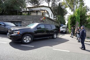 Ein schwarzes Auto biegt von einer Grundstückseinfahrt auf eine Straße ab, an der Seite stehen Sicherheitsleute