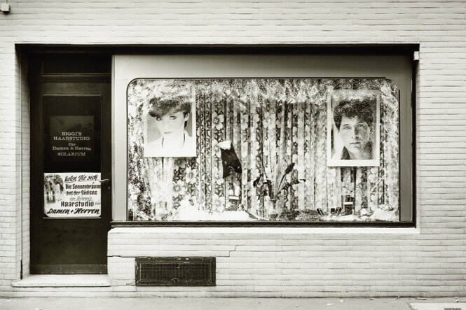 Sicht auf das Schaufenster eines Friseurladens in Köln 1980 mit den verblassten Fotografien von Frisurmodels vor zugezogenen Gardinen